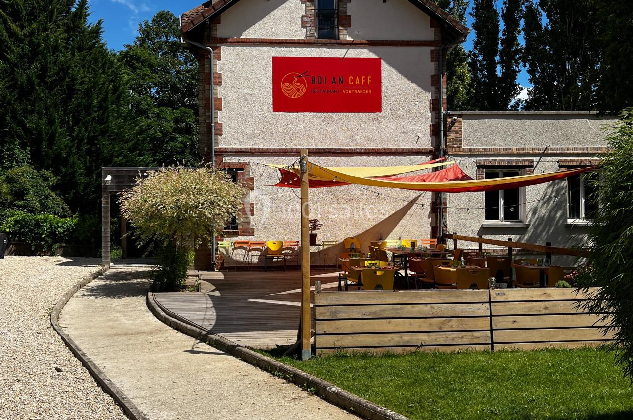 Façade d'un café avec terrasse extérieure aménagée, entourée de végétation sous un ciel bleu.