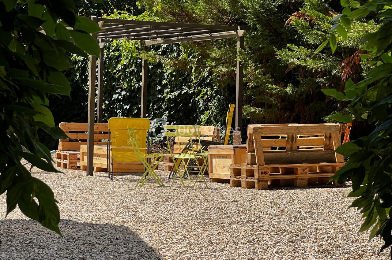 Espace extérieur avec salon de jardin en palettes, chaises jaunes et pergola, entouré de végétation.