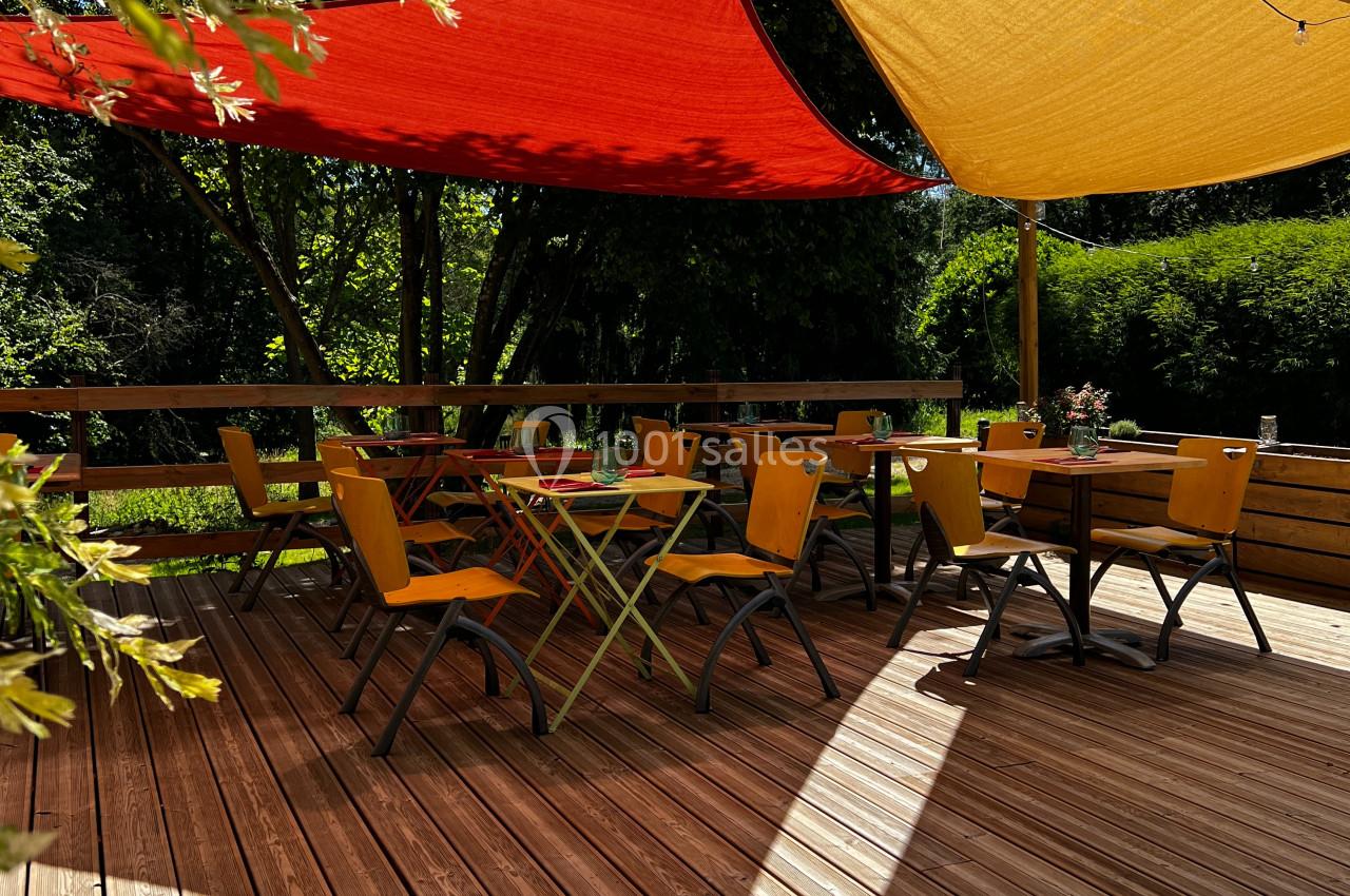 Terrasse en bois avec des tables et chaises jaunes, ombragée par des voiles rouges et jaunes, entourée de verdure.