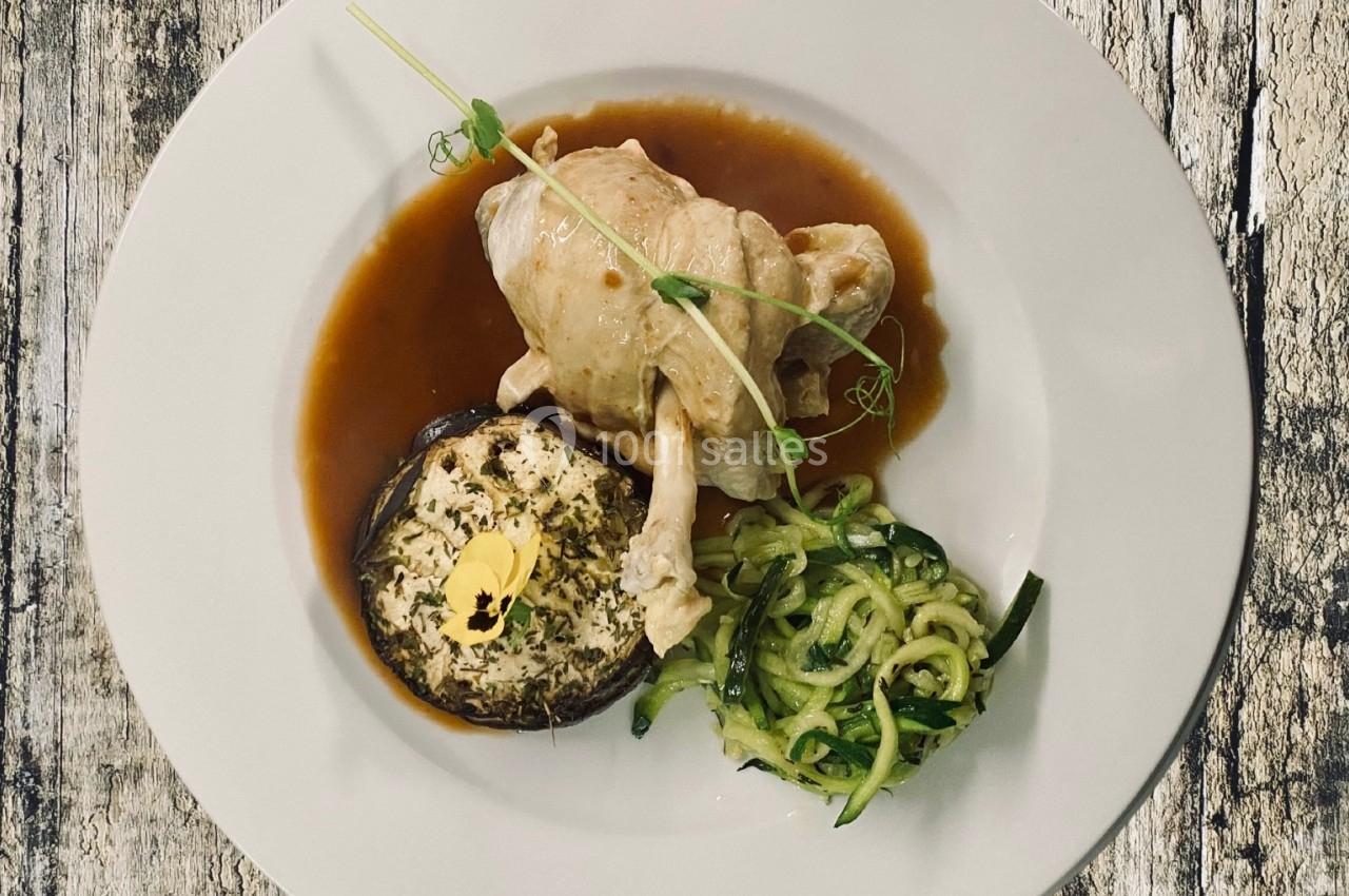 Assiette avec un suprême de volaille, une courgette spiralée, un champignon farci et une sauce brune, sur une table en bois.