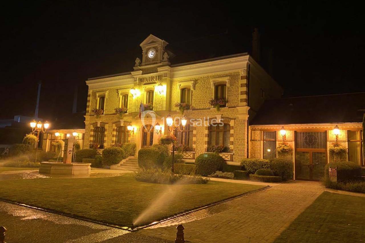 Bâtiment éclairé de nuit avec une horloge en façade, entouré de jardins et d'allées arrosées.