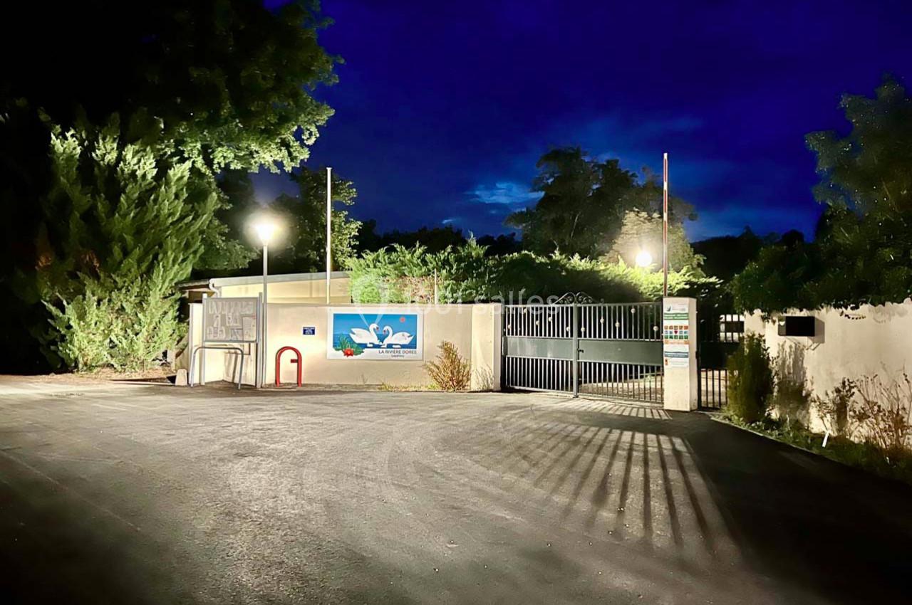Entrée d'un bâtiment avec portail fermé, éclairée par des lampadaires, entourée de végétation sous un ciel nocturne.