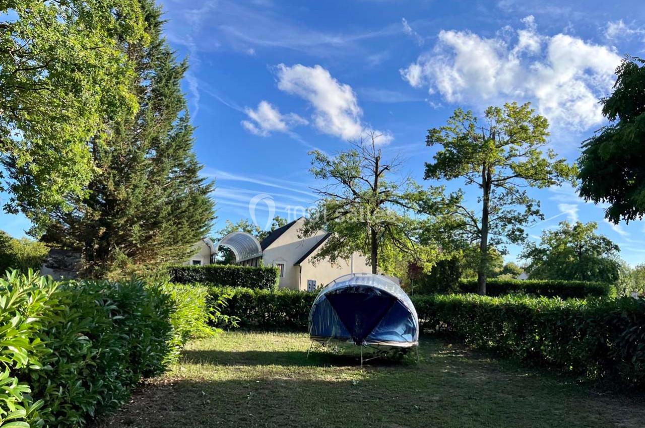 Tente bleue installée sur une pelouse entourée de haies et d'arbres, avec une maison blanche en arrière-plan.