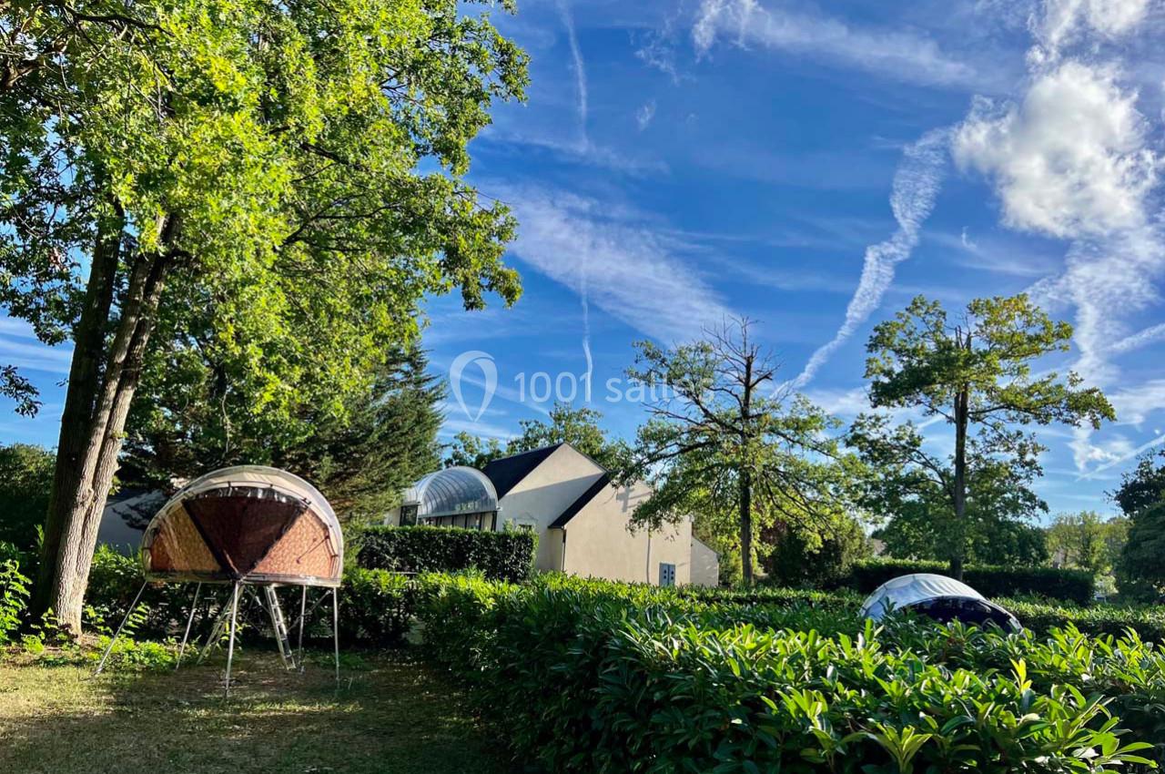 Deux tentes suspendues parmi des arbres dans un jardin ensoleillé, avec une maison en arrière-plan et un ciel bleu strié de…