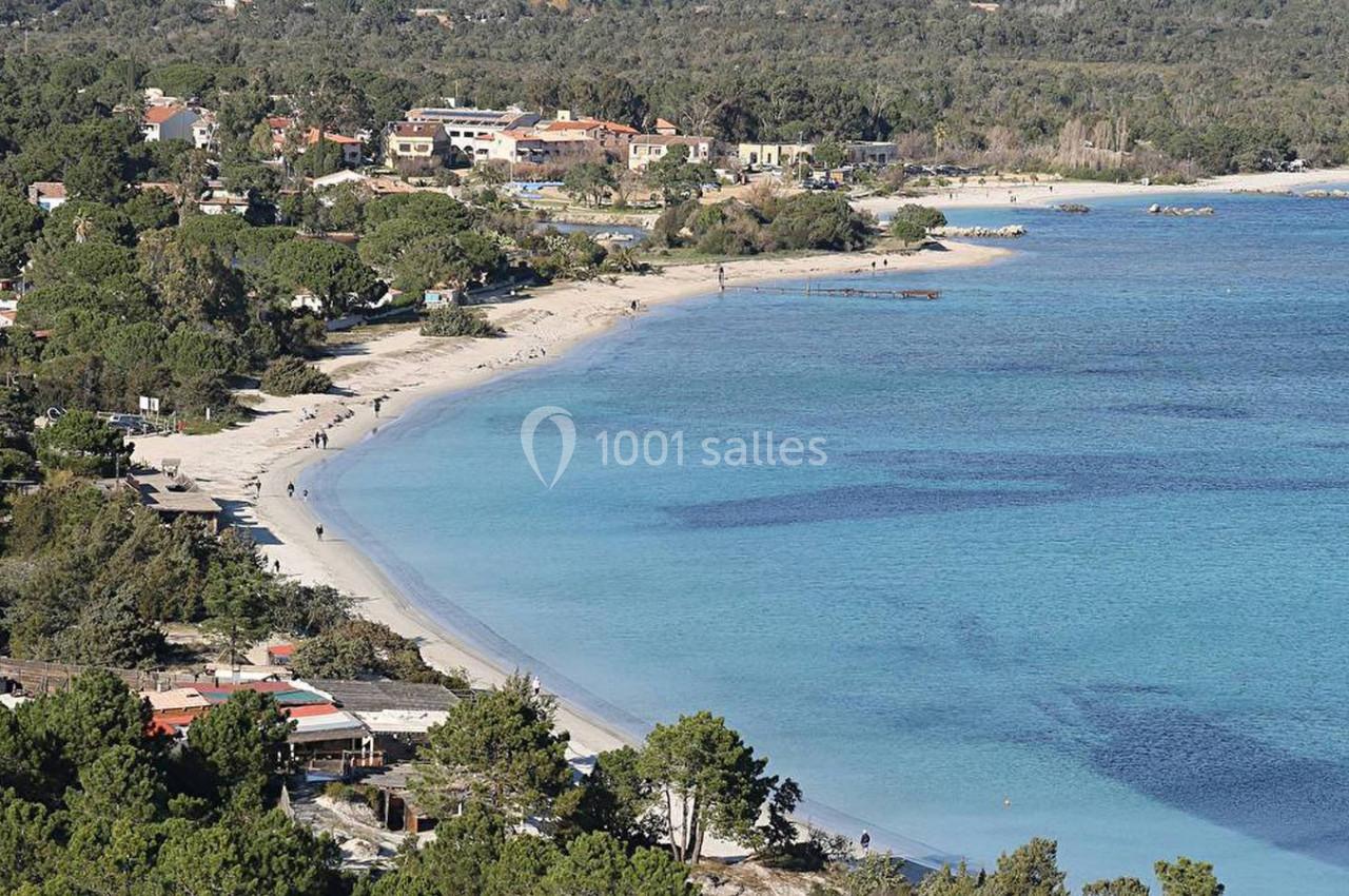 Vue aérienne d'une plage bordée de végétation et de bâtiments, avec une mer calme et des nuances de bleu.