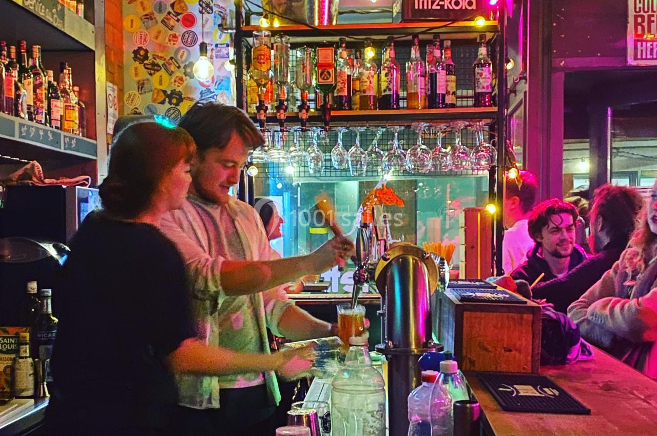 Un barman et une barmaid préparent des boissons dans un bar animé, entourés de bouteilles et de lumières colorées.