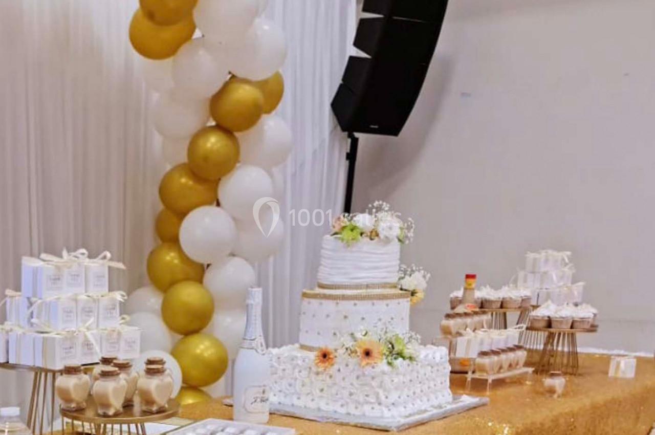 Table décorée pour une fête avec un gâteau blanc orné de fleurs, ballons dorés et blancs, et desserts variés.