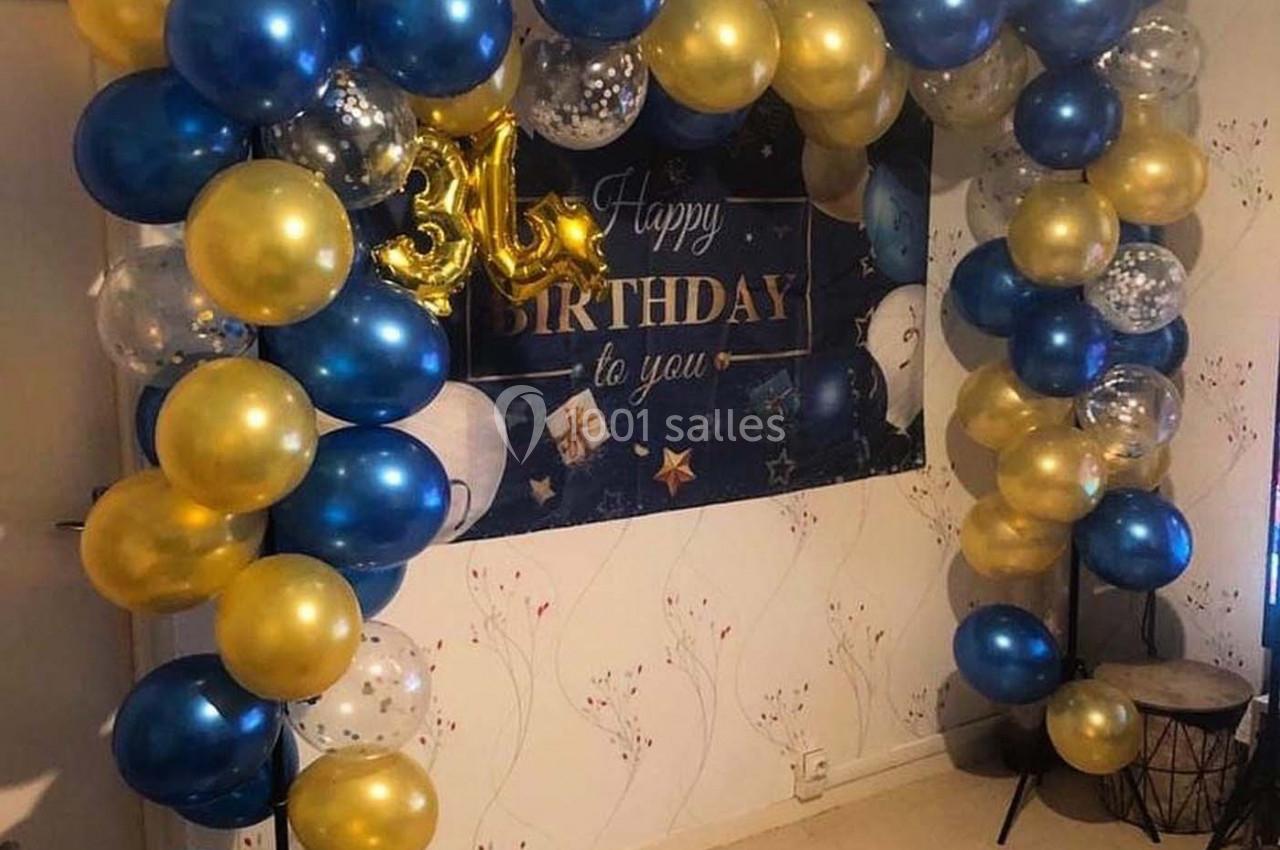 Arche de ballons bleus, dorés et transparents décorant une pièce avec une bannière d'anniversaire en arrière-plan.