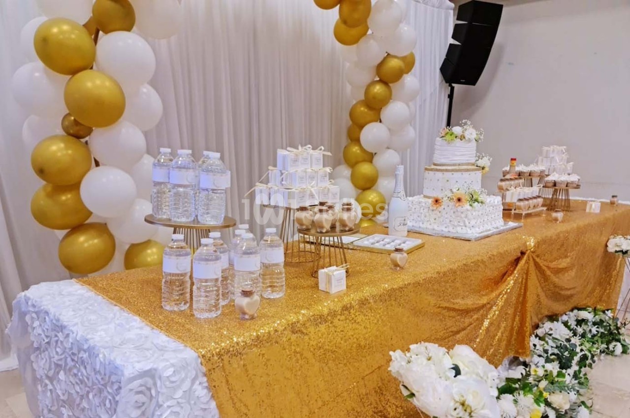 Table décorée pour une fête avec nappes dorées, ballons blancs et dorés, gâteaux, bouteilles d'eau et fleurs blanches.