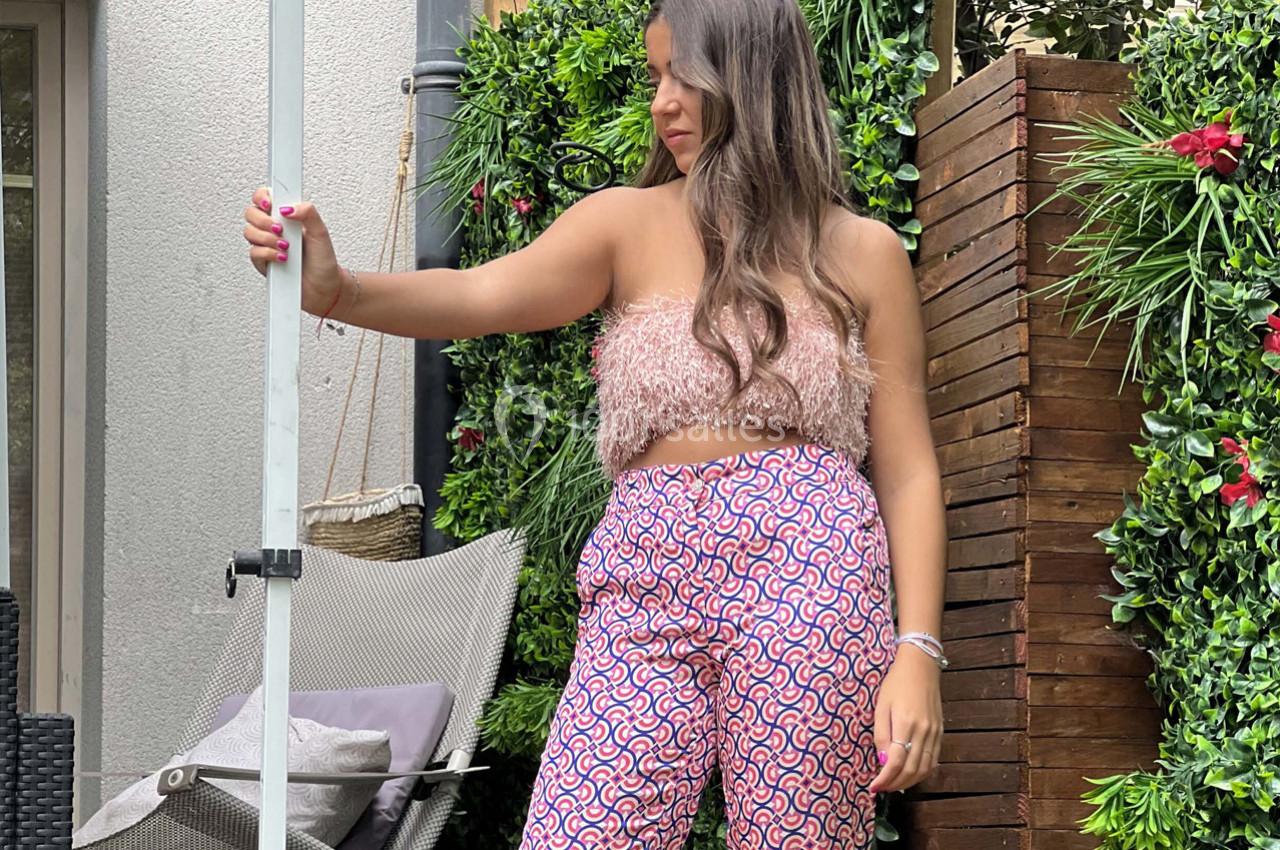 Femme debout dans un jardin, portant un haut texturé et un pantalon à motifs, tenant un poteau métallique.