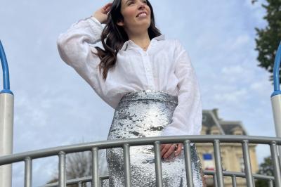 Femme souriante portant une jupe argentée scintillante et une chemise blanche, debout sur une structure métallique en…