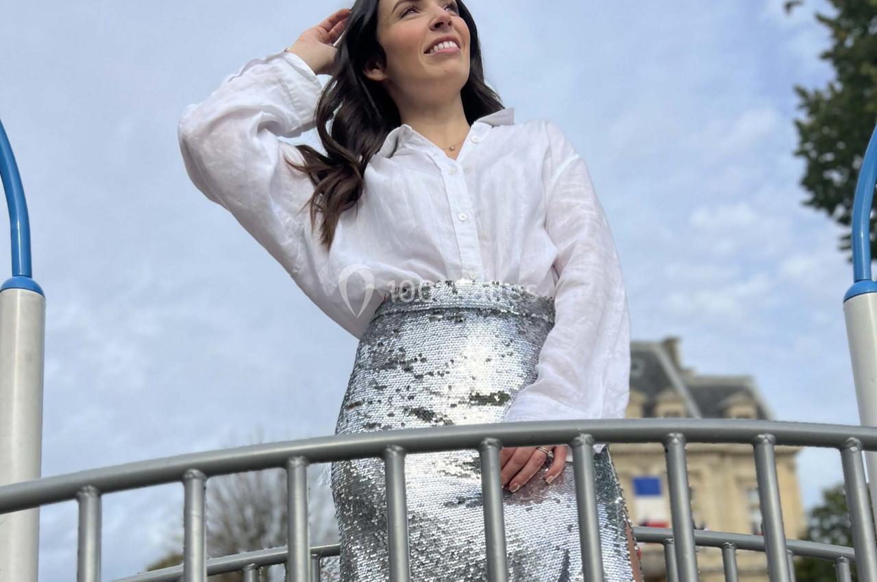 Femme souriante portant une jupe argentée scintillante et une chemise blanche, debout sur une structure métallique en…