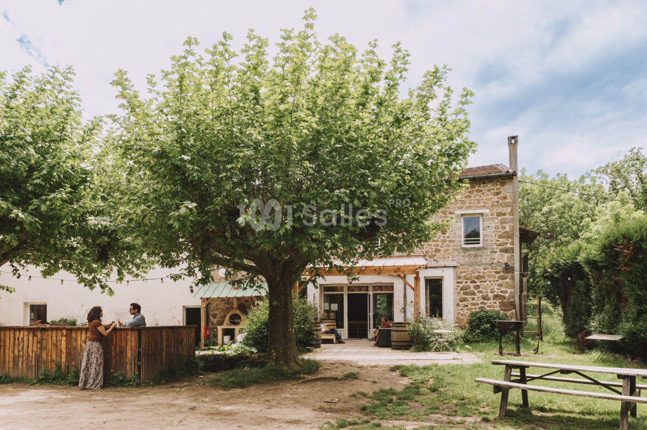 Maison en pierre entourée de verdure avec des arbres, une terrasse, des personnes discutant et une table de pique-nique.