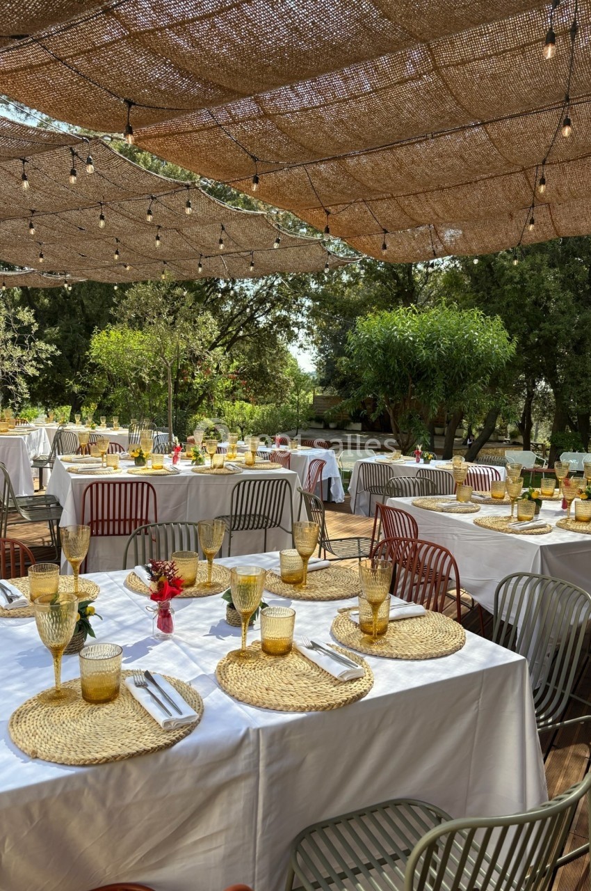 Tables dressées en extérieur avec nappes blanches, sets en osier, verres ambrés et guirlandes lumineuses sous une pergola.