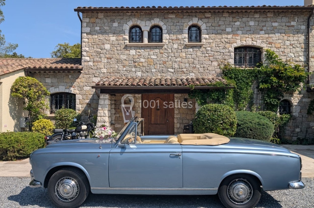 Voiture ancienne décapotable bleue garée devant une maison en pierre avec des volets et des plantes grimpantes.