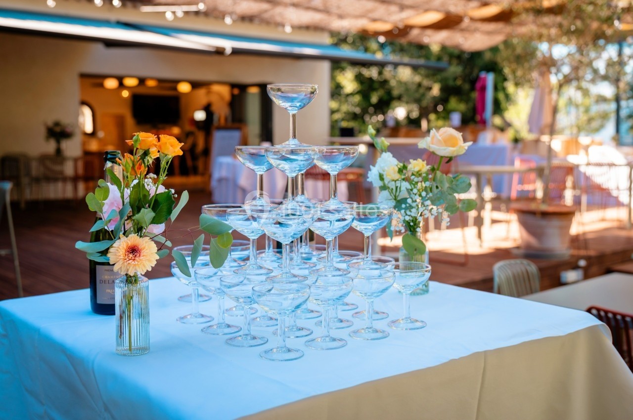 Pyramide de coupes à champagne sur une table blanche, entourée de bouquets de fleurs, en extérieur.