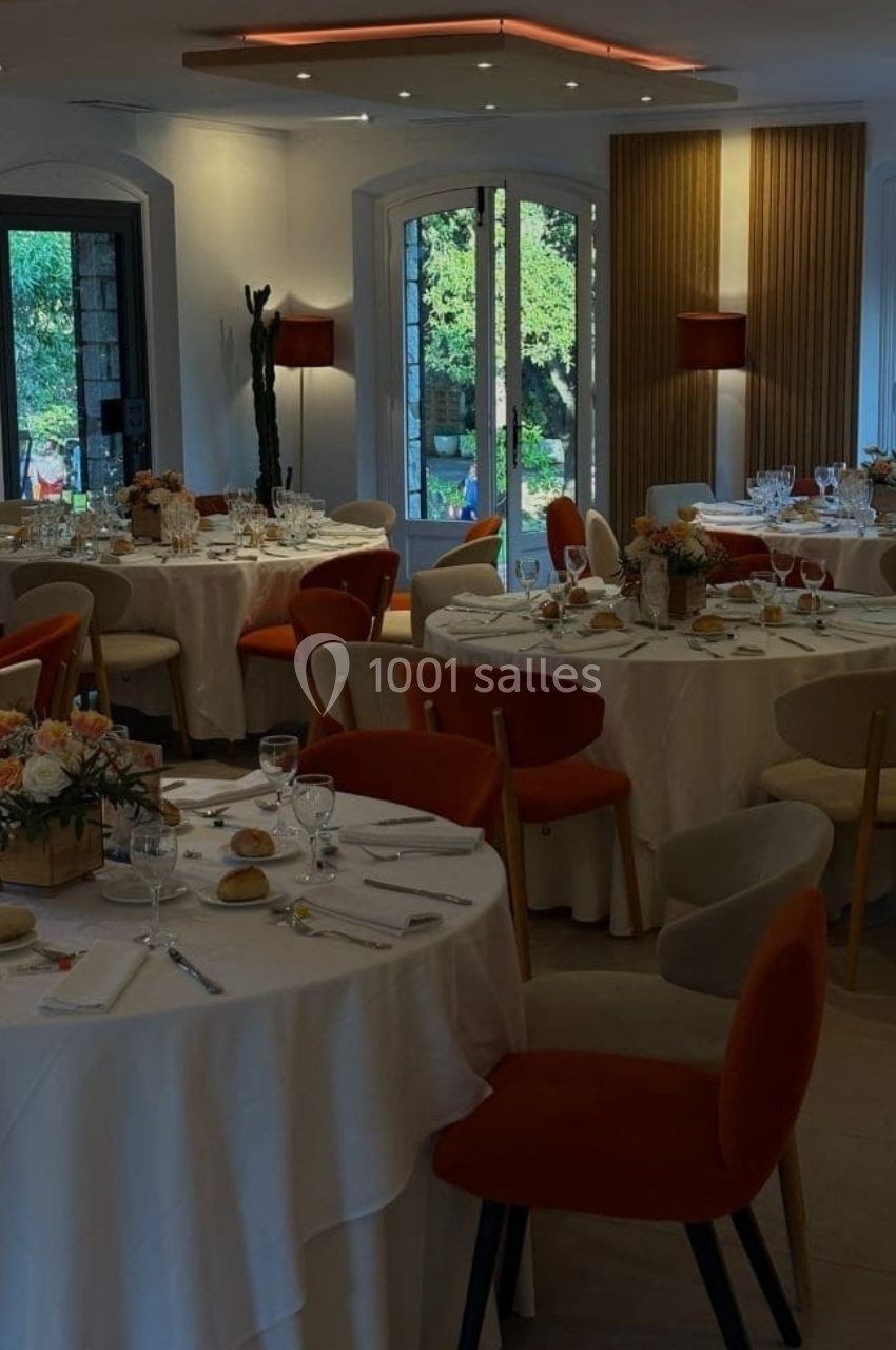 Salle de réception élégante avec tables rondes dressées, nappes blanches, chaises colorées et vue sur un jardin.