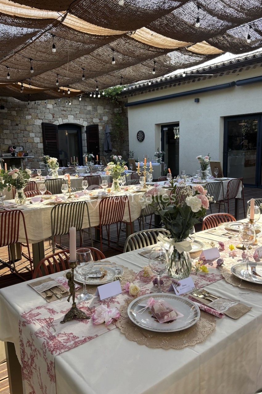 Tables décorées pour un repas en extérieur, avec nappes blanches, fleurs, bougies et vaisselle élégante sous une pergola.
