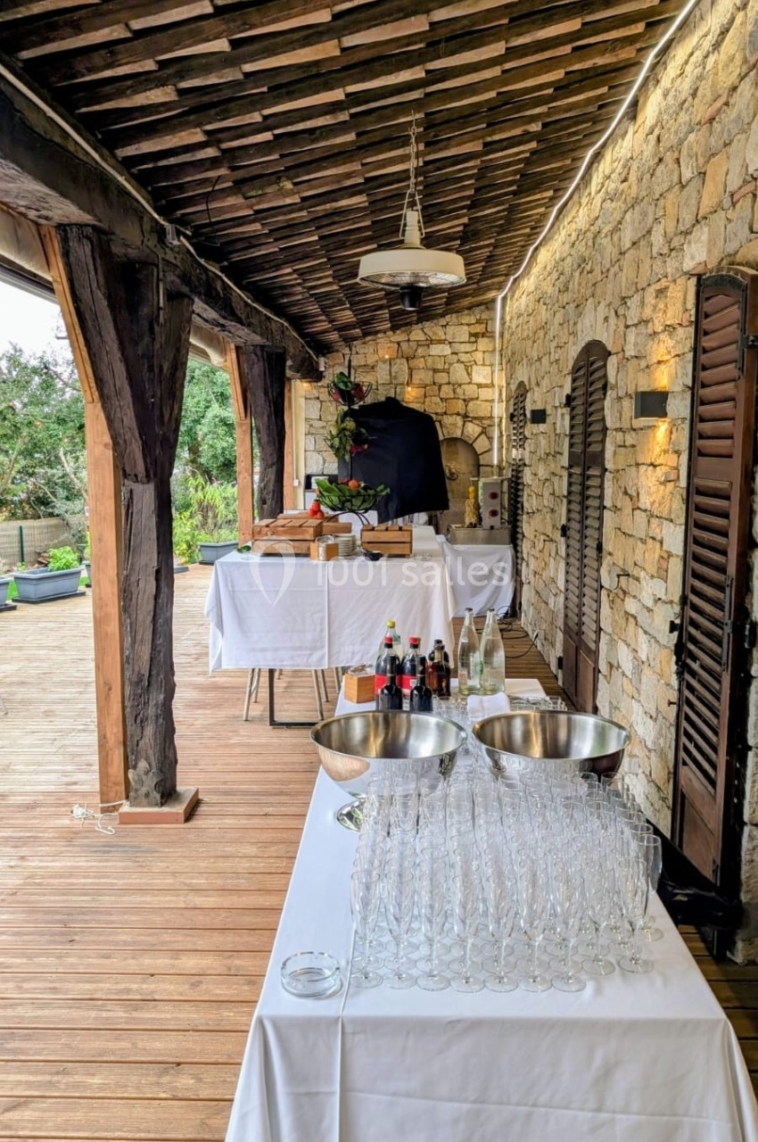 Table dressée avec verres, bouteilles et plats sous une terrasse en bois avec murs en pierre.