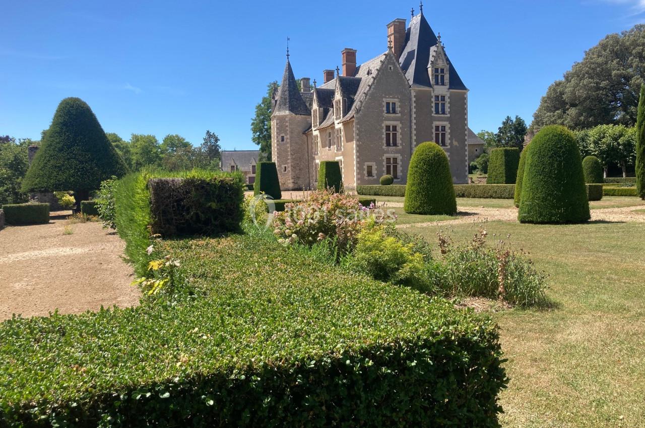 Château en pierre entouré de jardins avec haies taillées et arbres sculptés sous un ciel bleu.