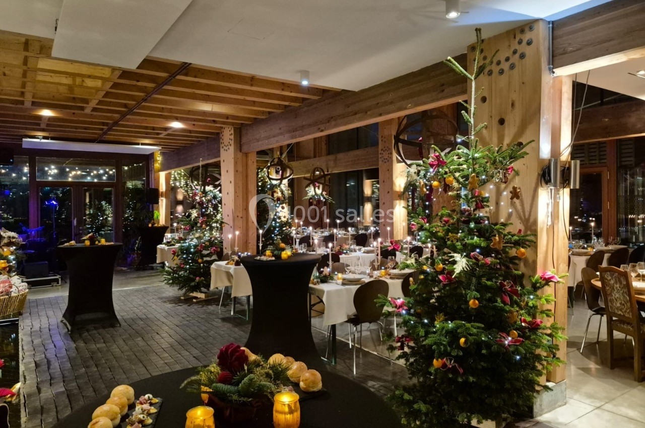 Salle décorée pour une fête avec sapins ornés, tables dressées et éclairage chaleureux.