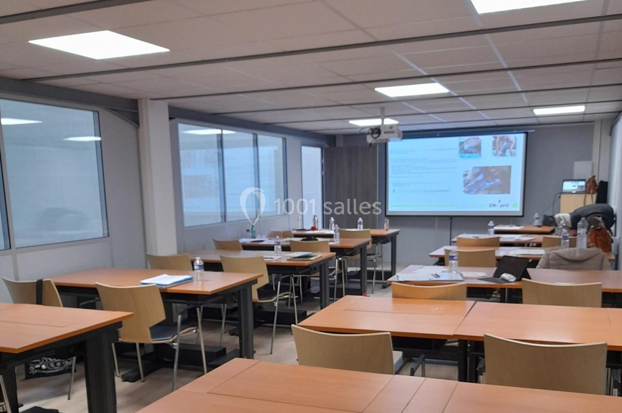 Salle de classe vide avec des tables en bois, des chaises, un projecteur allumé et des documents posés sur les tables.