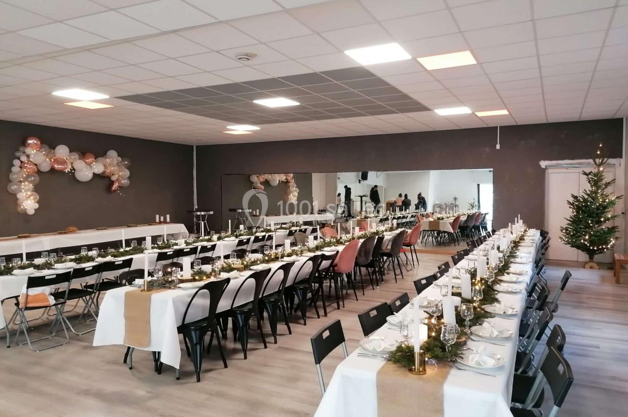 Salle décorée pour un événement avec de longues tables dressées, bougies, vaisselle et guirlandes lumineuses.