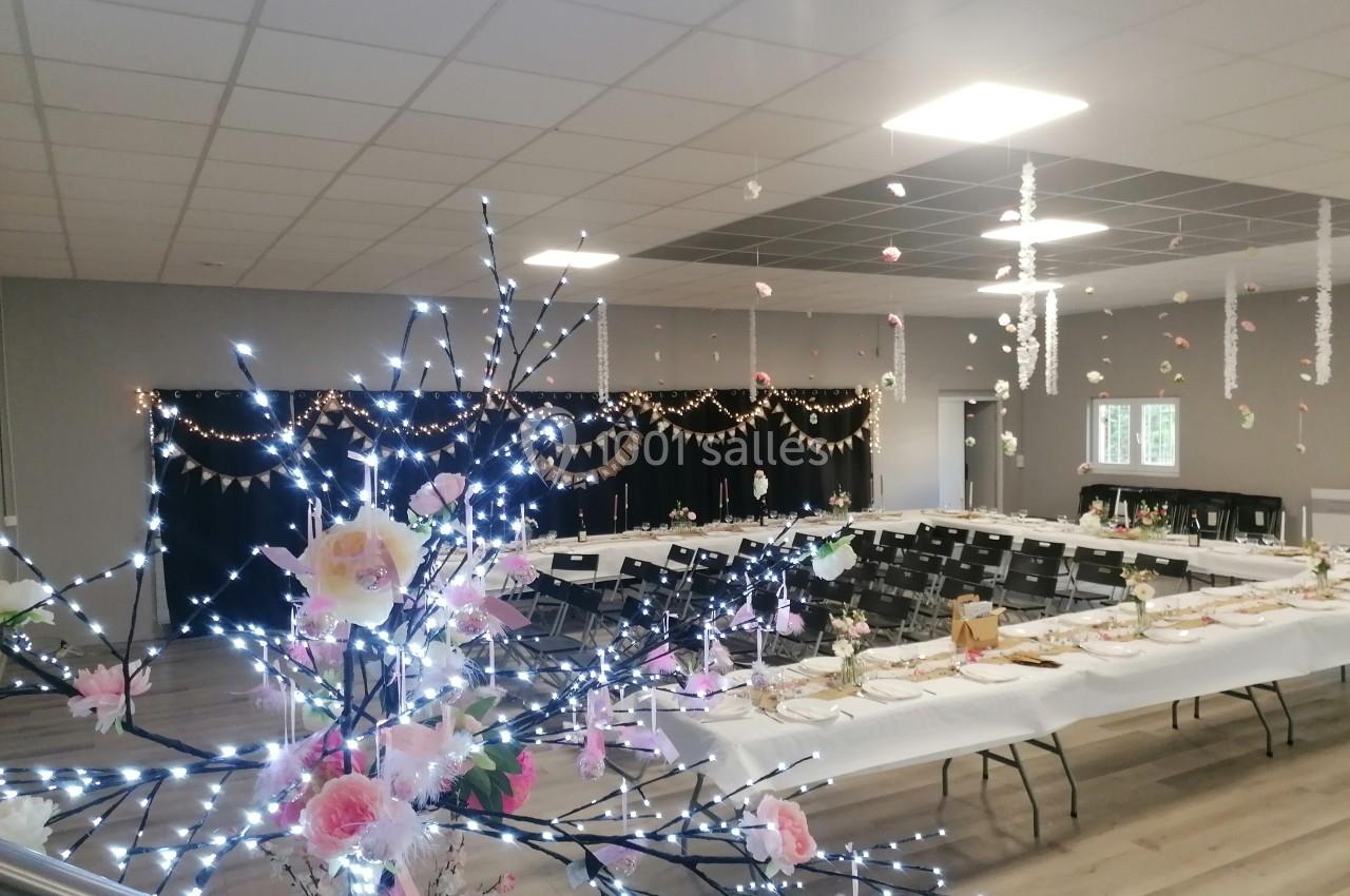 Salle décorée pour un événement avec tables dressées, guirlandes lumineuses, fleurs et ornements suspendus.