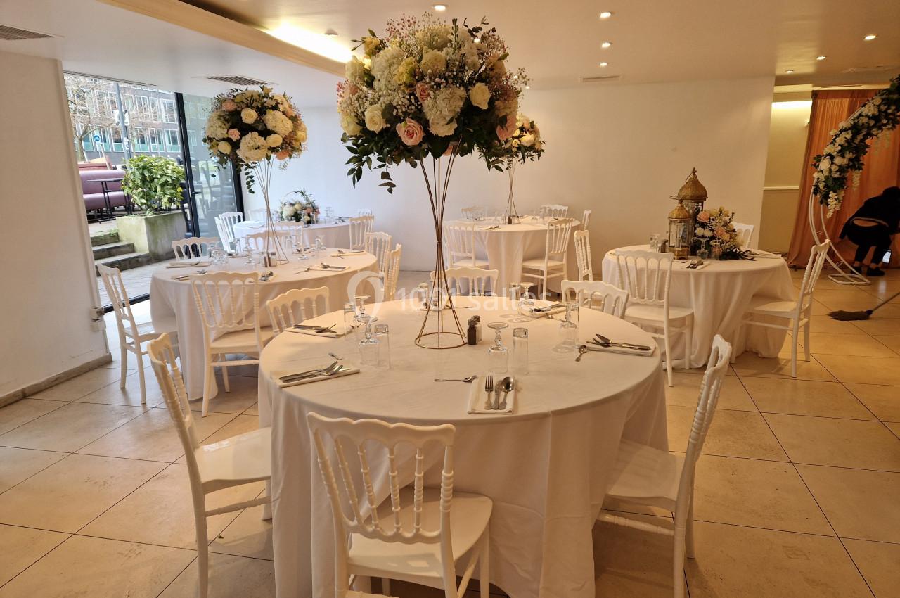 Salle de réception décorée avec des tables rondes, nappes blanches et centres de table fleuris sur pied.