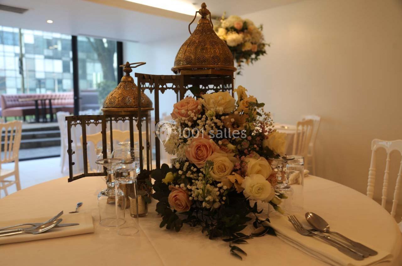 Centre de table avec lanternes dorées et bouquet de fleurs pastel, disposé sur une table dressée dans une salle lumineuse.
