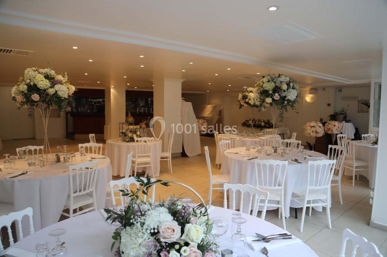Salle de réception lumineuse avec tables rondes décorées de fleurs blanches et roses, chaises blanches et éclairage doux.