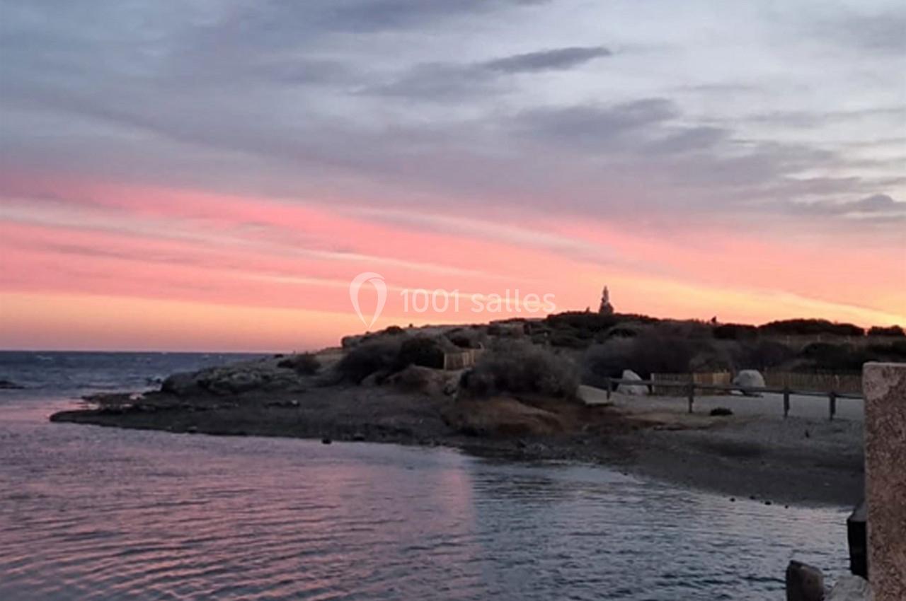 Coucher de soleil aux teintes roses et violettes sur une plage avec une petite colline et un bâtiment en arrière-plan.