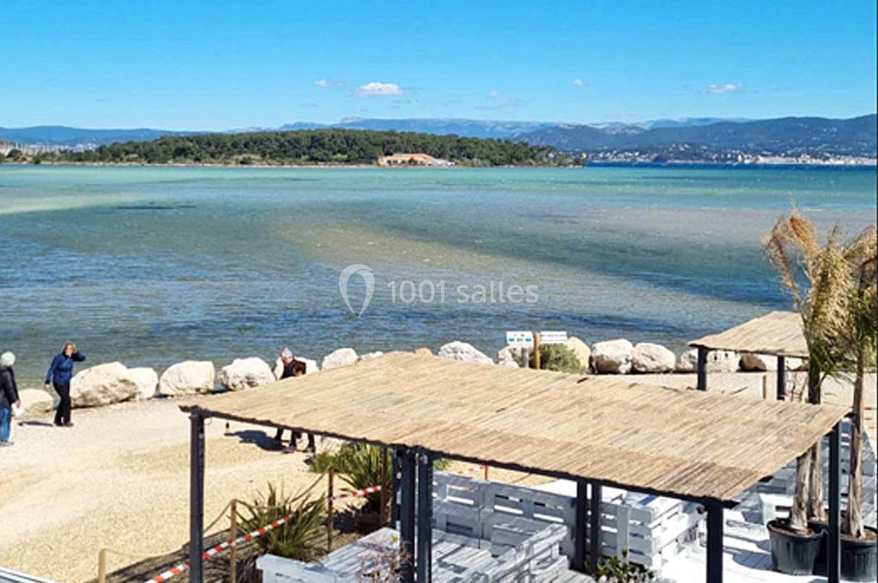 Vue d'une plage avec une terrasse ombragée, des promeneurs, une étendue d'eau et des collines à l'horizon.