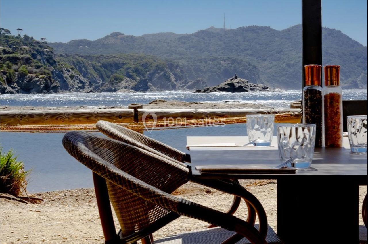 Chaises en osier et table dressée avec vue sur une plage, des collines et l'océan sous un ciel dégagé.