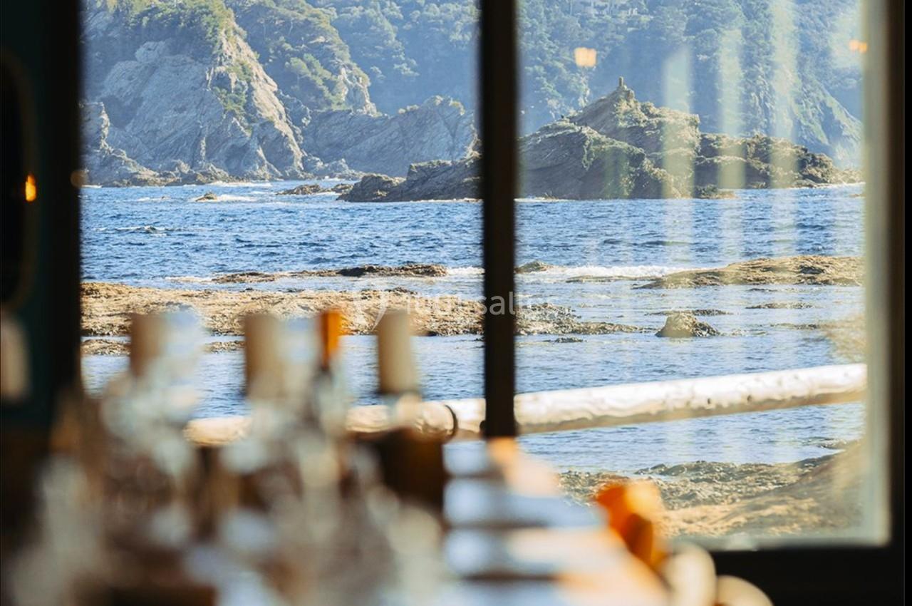 Vue sur la mer et les falaises à travers une fenêtre, avec une table dressée floutée au premier plan.