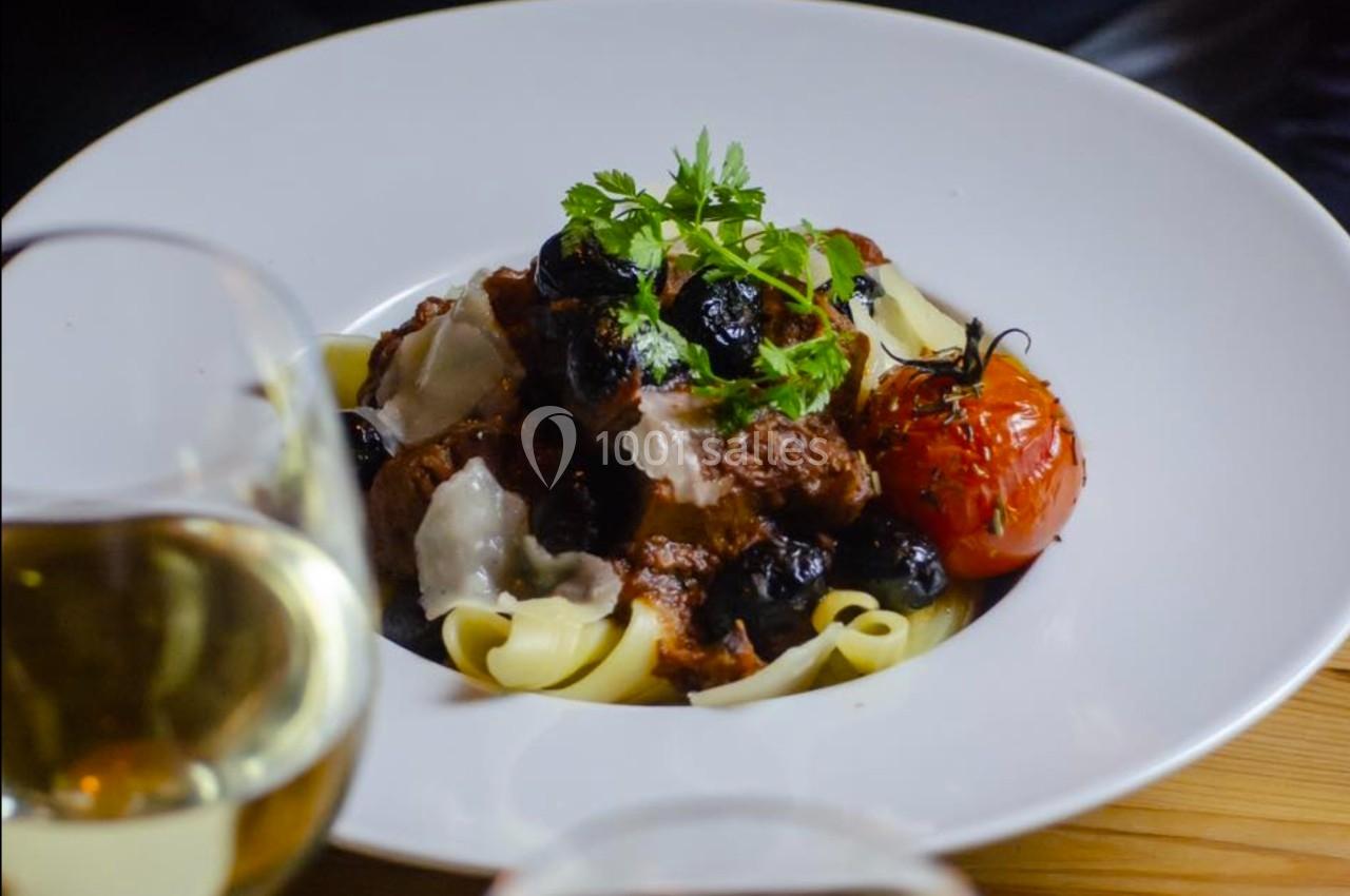 Assiette de pâtes garnie de viande en sauce, olives noires, tomate rôtie et herbes, servie avec du vin blanc.