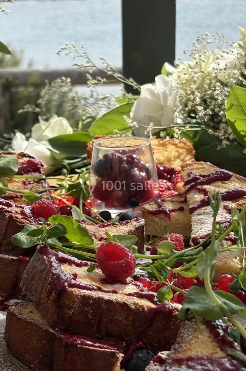 Tranches de brioche dorées garnies de fruits rouges, nappées de coulis, entourées de verdure et de fleurs, en extérieur.