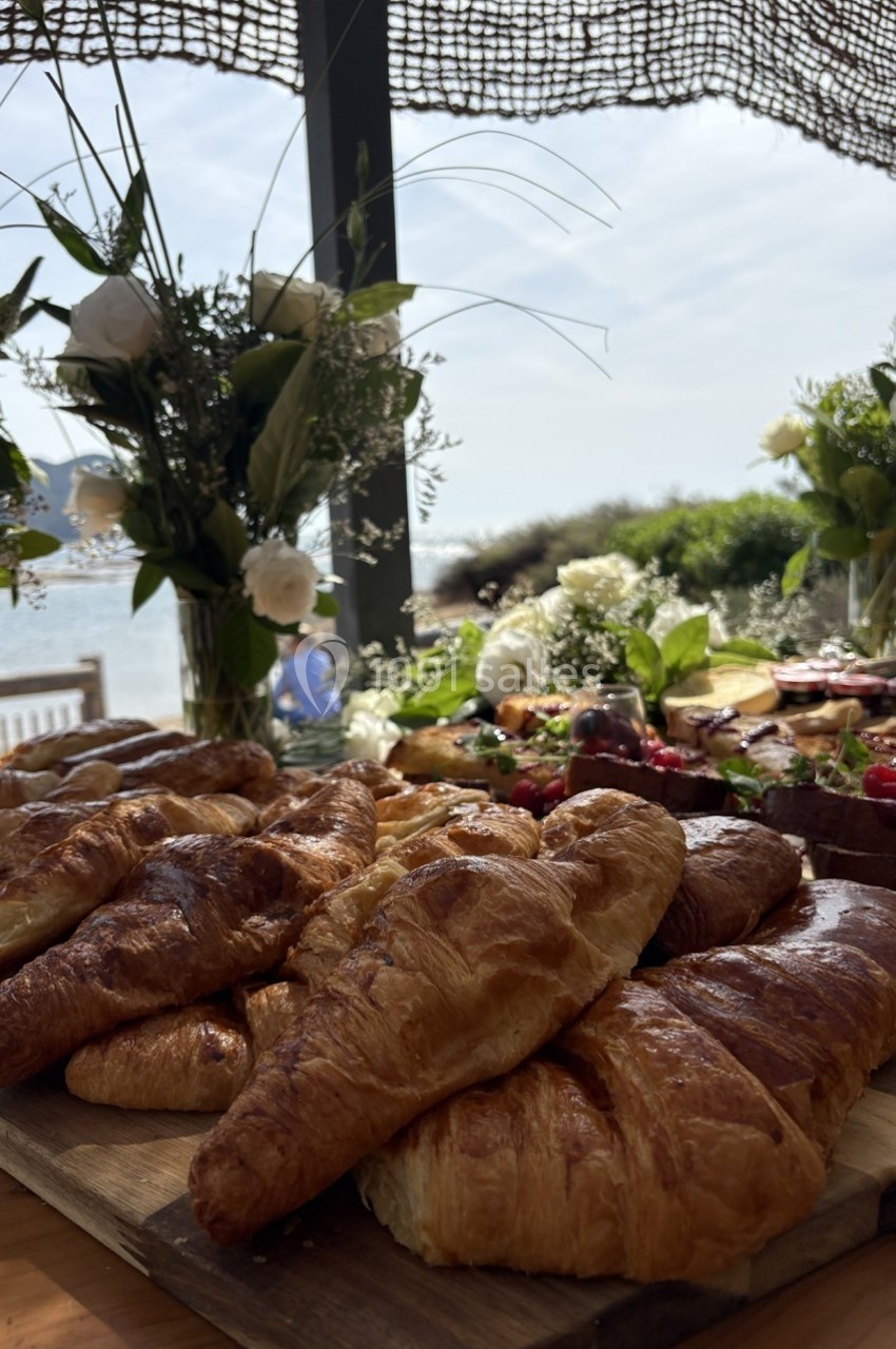 Plateau de croissants dorés disposés sur une table en bois, entourés de fleurs et d'autres mets, avec vue sur la mer.