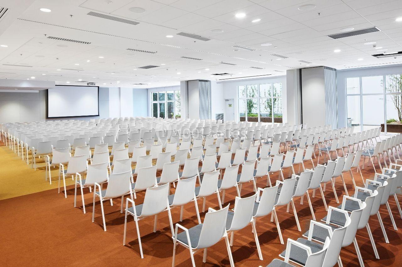 Salle de conférence lumineuse avec rangées de chaises blanches alignées face à un écran de projection.