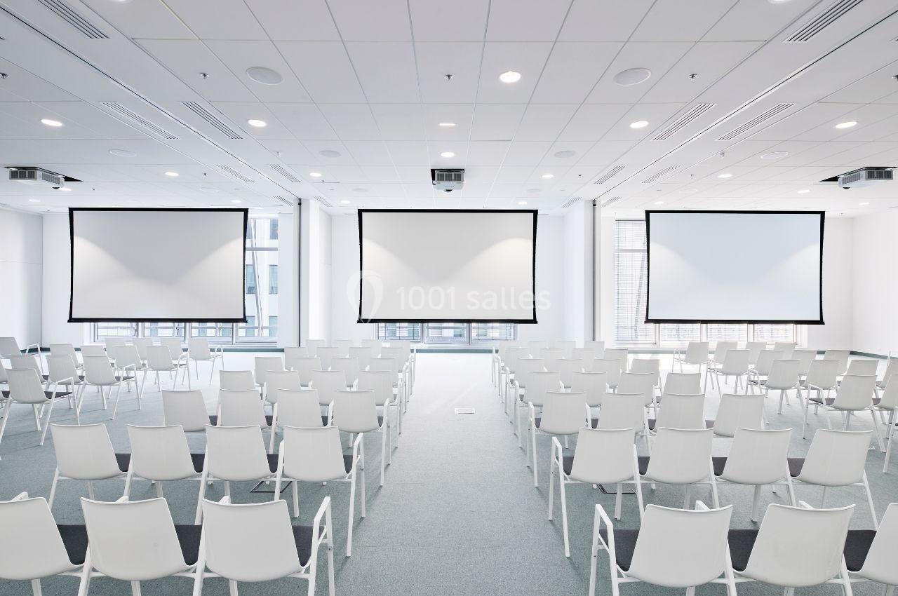 Salle de conférence lumineuse avec des rangées de chaises blanches et trois écrans de projection alignés.