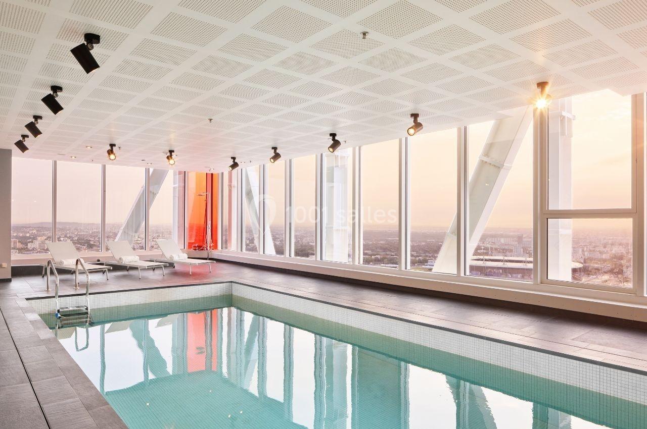 Piscine intérieure avec baies vitrées offrant une vue panoramique sur la ville au coucher du soleil.
