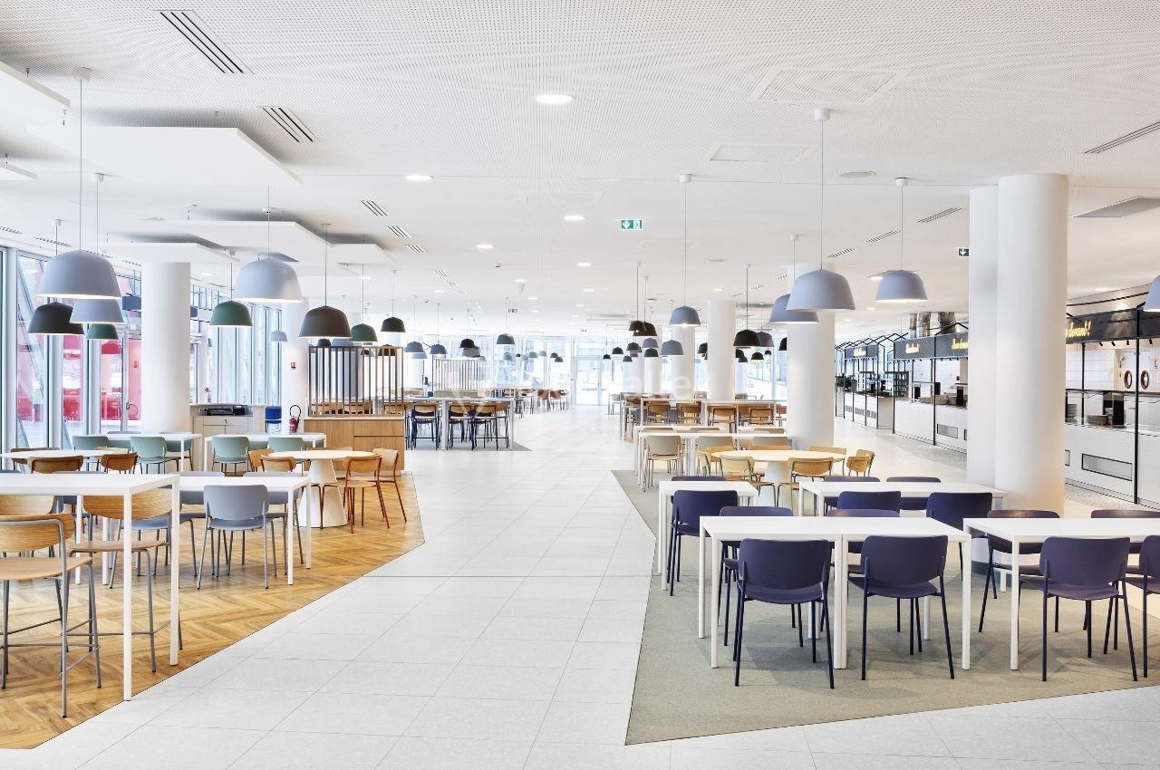 Salle lumineuse avec tables et chaises modernes, divisée en zones avec différents types de sols et éclairages suspendus.