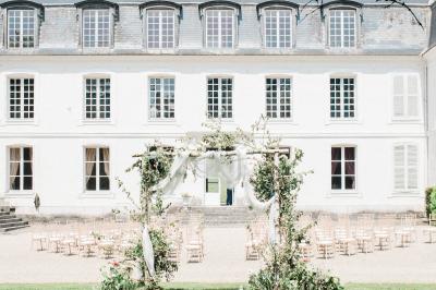 Femme en veste rouge lisant un texte sur un pupitre décoratif, devant un mur en pierre.