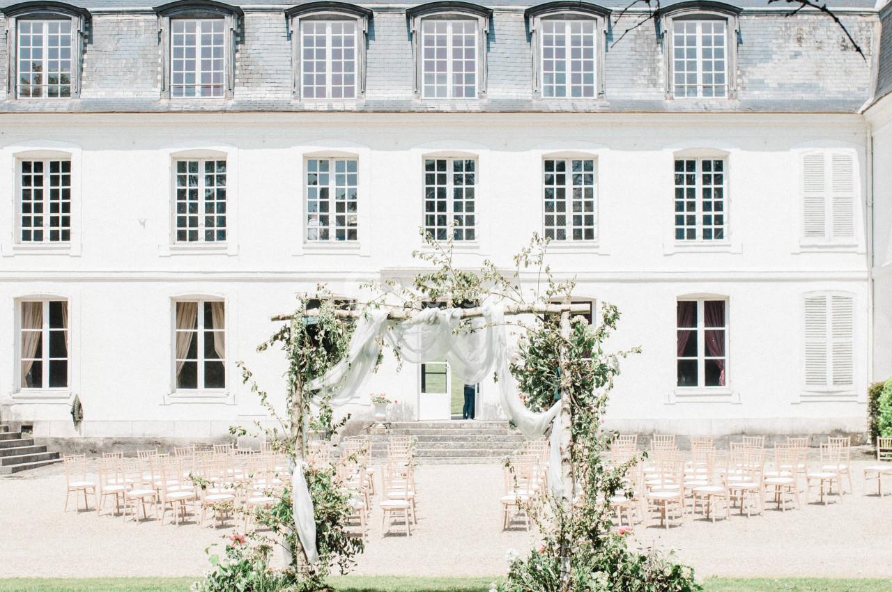 Façade d'un bâtiment blanc de style classique avec une arche décorée et des chaises disposées en extérieur.