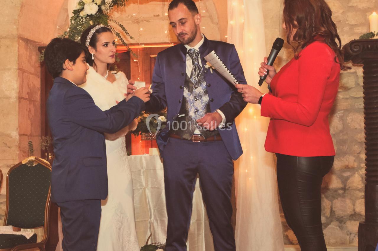 Un couple en tenue de mariage échange des vœux devant une femme avec un micro et un jeune garçon dans une salle en pierre.
