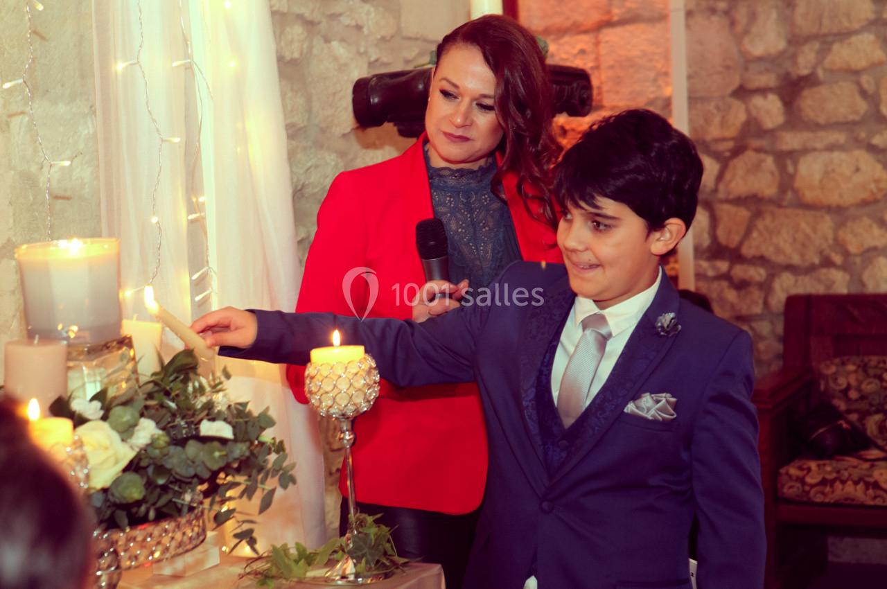 Un garçon en costume allume une bougie lors d'une cérémonie, accompagné d'une femme tenant un micro.