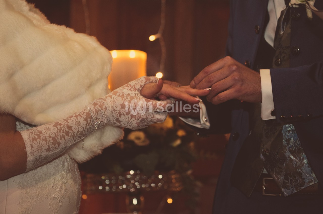 Un marié passe une alliance au doigt de la mariée portant des gants en dentelle, devant une bougie allumée.