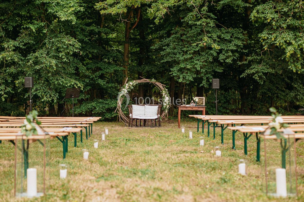 Chaises et arche décorée en bois installées dans une clairière, entourées de bancs et de bougies blanches au sol.