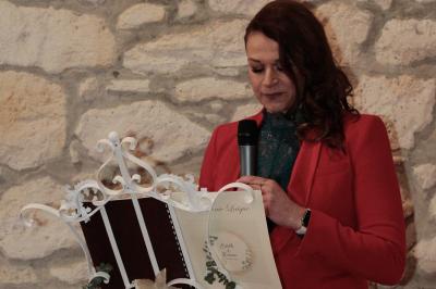 Femme en veste rouge lisant un texte sur un pupitre décoratif, devant un mur en pierre.