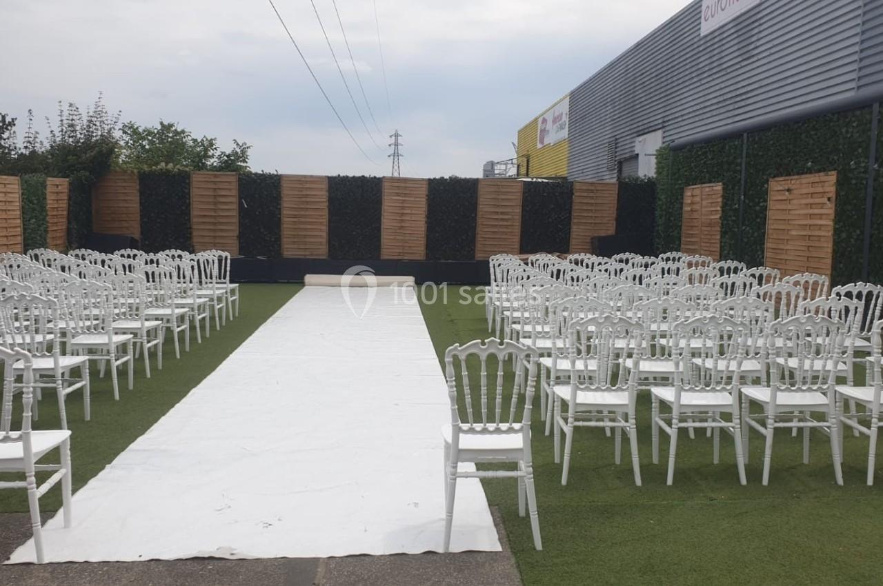 Allée centrale bordée de chaises blanches alignées, installée en extérieur sur une pelouse synthétique.